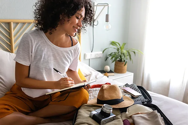 Woman sitting on bed writing packing list in a notebook